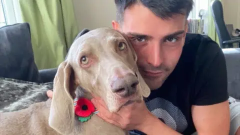 Jordan Adams Jasmine, a silver‑grey Weimaraner sits calmly indoors, wearing a red poppy collar. Jordan Adams is gently holding the dog’s face.