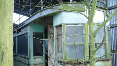 Brighton Tram 53 Society A rotting tram car in a metal barn. It is covered in green fungus with a tree growing from its front