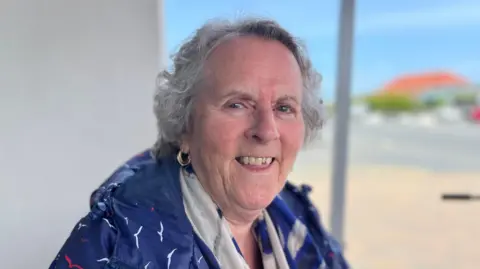 A lady with short grey hair smiling towards camera, wearing a blue jacket. 