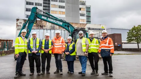 Stockton Council Members of the Thornaby Town Deal Board (L-R): Matt Vickers MP, councillor Ian Dalgarno, Mark White CBE DL, councillor Sylvia Walmsley, councillor Richard Eglington, councillor Nathan Gale and Contractor MGL Group’s Joint Chief Executive Mark Davison senior, standing outside a derelict tall hotel, with a crane behind them.