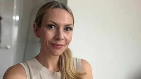 Aimee Bradley Foto de cabeza y hombros de una mujer caucásica con cabello largo y rubio recogido en una cola de caballo sobre su hombro izquierdo y sonriendo a la cámara. Lleva un chaleco beige. 