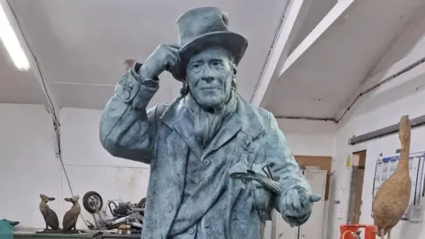 Sebastien Boyesen Design The statue of Jimmy Strength is stood in a workshop. He has a tall hat on his head and a violin under his arm. He is a dark grey colour.
