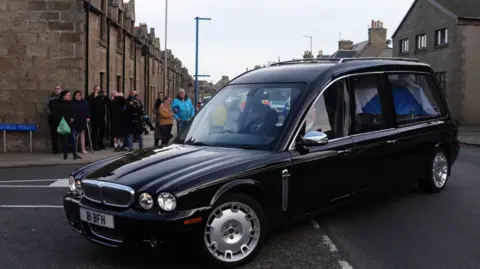 PA Media Black hearse turns corner of street with small crowd watching from pavement in front of historic row of houses.