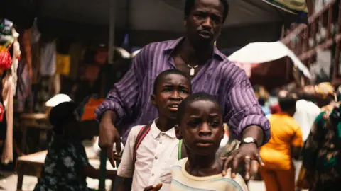 MUBI Actor Sope Dirisu walks down a dusty road whole holding the hands of young actors Godwin Chiemerie Egbo and Chibuike Marvellous Egbo. Sope is wearing a purple striped shirt and jeans. The boys are wearing jeans and T-shirts. 