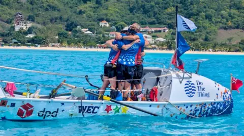 Four people on a boat hugging one another in blue water. Behind them is a beach with green trees and plants and a few white and terracotta buildings.