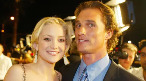 Kate Hudson and Matthew McConaughey standing together and smiling in formal dress, with red carpet activity behind them