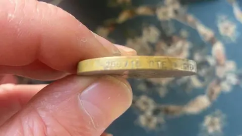 Steve Knighton A close-up on some fingers holding a metal medal with some letters and numbers engraved on its side