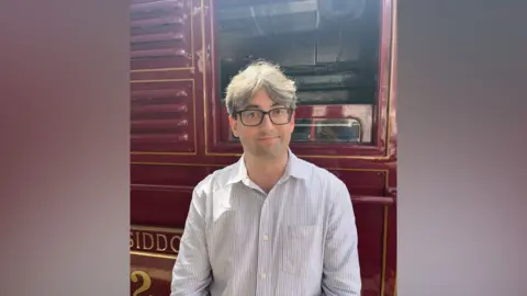 A man in light grey shirt with glasses and grey hair stands in front of a vintage locomotive.