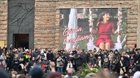 AFP via Getty Images A large crowd of people outside the Basilica of Santa Giustina. On the wall of the basilica is a large poster several metres high showing Giulia Cecchettin in a red dress sitting on a swing. 