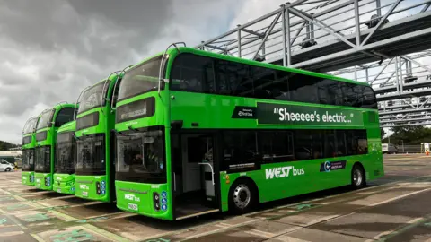 Four lime green double decker buses lined up next to each other at a bus depot. One of the buses says "Sheeeee's electric..." in white writing.