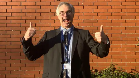 James Suther pulling a funny face with two thumbs up. He is wearing a star wars tie and a black blazer with a blue shirt. He is stood against a brick wall. 