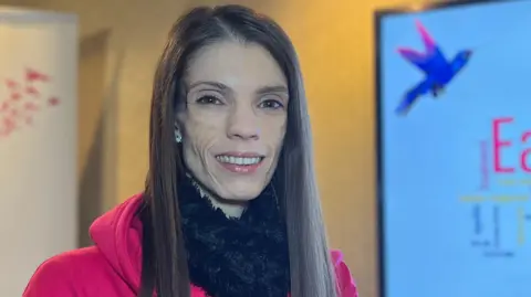 Amy is in a bright red hoodie stands indoors between a tall JEDS banner featuring a stylized bird and a presentation screen displaying a colorful word cloud related to eating disorders.