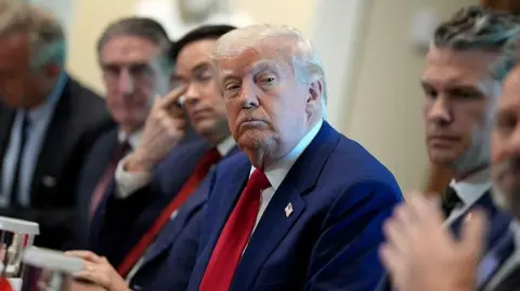 Trump is shown in a blue suit and red tie, with a sceptical look on his face, during a cabinet meeting, seated next to Secretary of State Marco Rubio and Secretary of Defence Pete Hegseth 