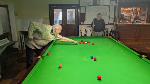 David Spereall/BBC Two gentlemen play snooker in a bar area. One is crouching over the table ready to play a shot, while the other stands and watches.