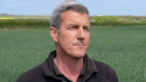 Farmer Rodd Smith looking side on with one his fields in the background. He has short black and grey hair and is wearing a brown shirt. He has a worried expression. 