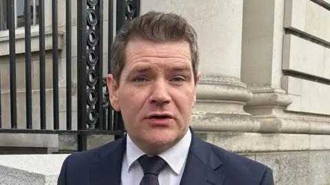 PA Media Peter Burke is standing outside government buildings in Dublin, he is wearing a navy suit with a white shirt and navy tie. He has short light hair with white patches. 
