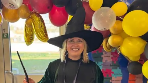 Elliot Deady/BBC A woman with shoulder-length blonde hair wearing a green cloak, black dress, and witch's' hat while holding a wand. She is smiling straight at the camera and standing in front of a balloon arch.