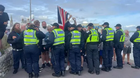 Seven police officers stand in a line in front of a group of men, one of whom is holding a Union Jack.