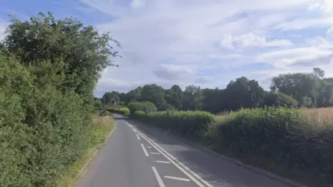 A view down the A4111. There are large hedgerows on either side of the route with a national speed sign on the left side of the road.