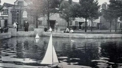 Port Sunlight Village Trust A black and white shot of the pond. There are children lining the sides, some with fishing lines. There are several model boats floating on the water.