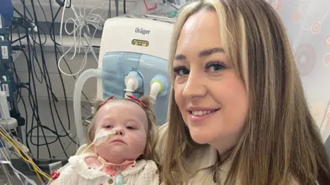 Poppy's mother, Becca, holds her in her arms. They are sitting in a hospital ward and medical equipment is behind them. Poppy has blonde hair and a tube is running from her nose.