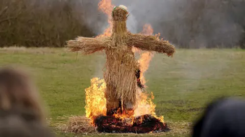 PA Media A wooden figure of a bear is in the middle of a field and is on fire. There are trees in the background. 
