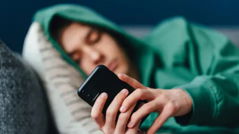 A stock image shows a teenage boy resting his head on a cushion on a sofa as he looks at his phone. His face is blurred out.