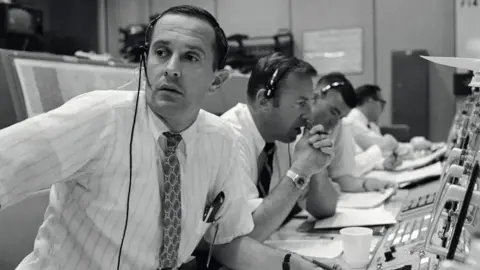 Imagen en blanco y negro de la NASA de Charlie Duke, junto con Jim Lovell y Fred Haise en la NASA Mission Control, durante la misión Apollo 11. Todos están vestidos con camisas y corbatas y usan auriculares, ya que se sientan en una larga línea frente a paneles de control y pantallas.
