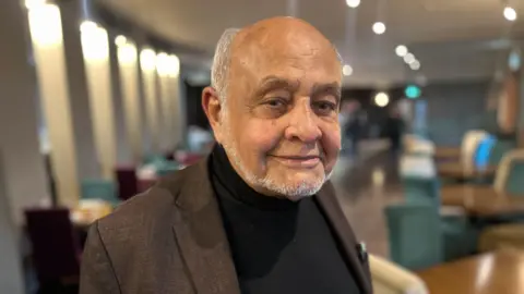 BBC news Cecil Balmond looking to the camera smiling wearing a black polo neck and a brown blazer. He is bald with some short grey hair to the side of his head and has a short grey beard.