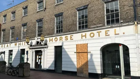 The front of the Great White Horse Hotel on the main high street in Ipswich. It is a white wood-panelled building, with the lettering in gold.