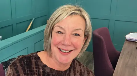 Grace Wood/BBC A woman with short blonde hair smiles at the camera. She is wearing a brown top and sitting in an office.