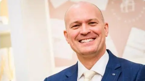 Tylers Department Store A smiling man wearing a blue suit and cream coloured tie. 