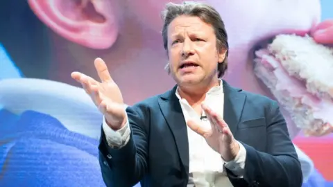 PA Media Chef Jamie Oliver at a public speaking event, gesticulating with his hands, in front of a screen showing a child eating a sandwich