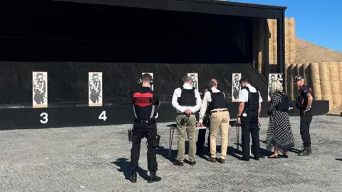 Five men and a woman all wearing bulletproof vests have their backs to the camera as they look at seven targets with men on them. Three of the targets are labelled 2, 3 and 4.