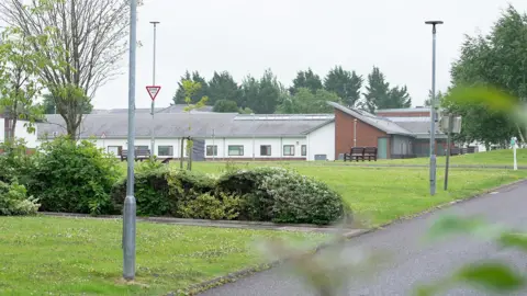 Muckamore Abbey hospital - a lowrise white and brick building - in the background with grass and shrubbery in the foreground, with a path alongside.