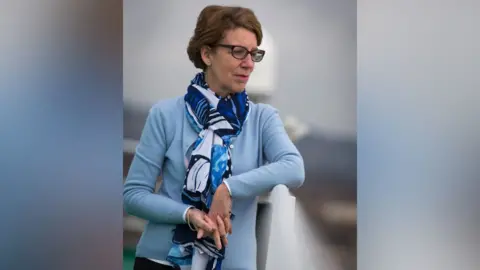 A woman with short brown hair and glasses leans on a white railing and looks to the side whimsically. She wears a light blue cardigan and a blue patterned scarf.