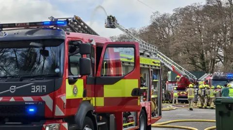 A fire engine in red and yellow is close on the left, and there are lots of crews in the background on the right. Above is an extended platform with two firefighters using a hose that is jetting out. There are trees behind to the right.
