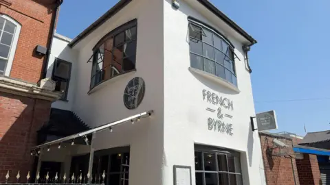 A white section of a building with bay windows on the ground and first floors. There is black lettering indicating the business occupying the building is French & Byrne. The building is set against a blue sky.