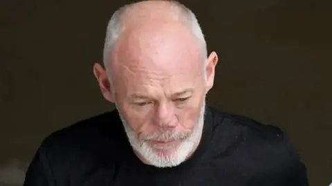 Andrew Pender is looking towards the ground, he is semi-bald with short grey hair and a great beard and moustache. He is wearing a black tee-shirt