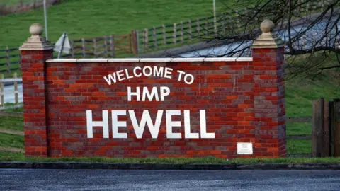 A red brick wall at the side of a road, saying 'Welcome to HMP Hewell' in white lettering. Wooden gates and grass are at the side of the road.