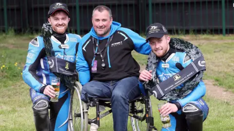 Isle of Man TT Races Ryan is on the left in blue and black leathers kneeling. Nick Crowe is in a wheelchair in the middle wearing the blue and black colours on his hoodie, and Callum kneeling to the right also in blue and black leathers. 