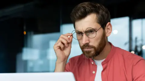 A man wearing a red shirt looks at a laptop screen, with a puzzled expressed on his face.