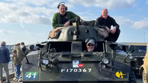 Three men sat on and in a small military tank. One man is peering out the window, while two men are sat on the turret.