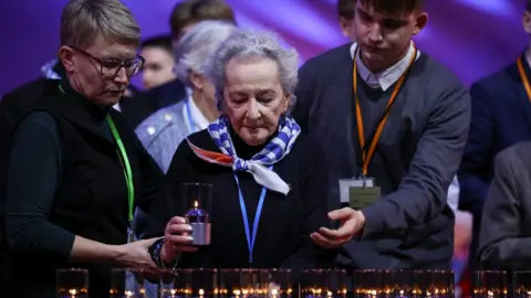 An elderly female holocaust survivor, helped by a younger woman and a young man, places a candle with during the commemoration