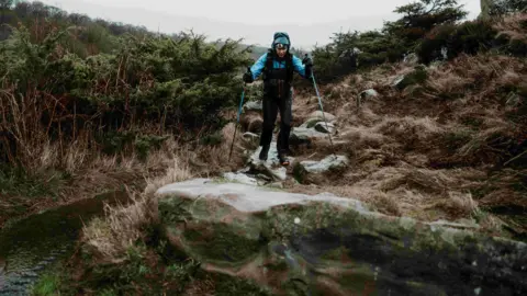 Clorroe Cam / Montane Winter Spine Race Runner Anna Troup on the Montane Winter Spine Race. She is wearing a blue waterproof jacket and a blue buff. She has a fell running backpack on and is holding blue running poles.