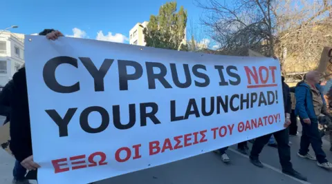 Jessica Parker/BBC People hold a white banner that reads: "Cyprus Is Not Your Launchpad!"