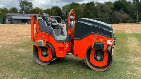 A large orange and black cricket pitch roller parked on a cricket lawn. 