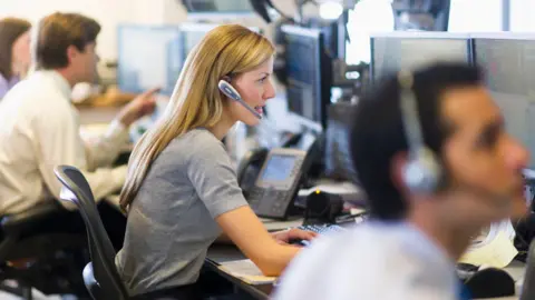 Getty Images Female trader with long blonde hair wearing a grey T-shirt and a phone headset, sitting at her desk looking at her screens, with other traders sitting to her left and right