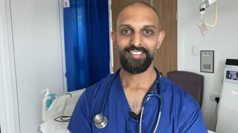 Ankur Gupta-Wright, one of the HCID Lead Consultants at Southmead Hospital. He has a black beard and short close cropped hair. He is wearing blue scrubs and is in a hospital.