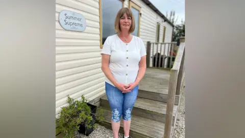 BBC Mandy Dossett, a woman in her 50s, is standing in front of her mobile home. Her brown hair is in a shoulder-length bob and she wearing a short-sleeved white blouse and denim capri trousers.